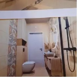 Jasna łazienka z beżowymi płytkami imitującymi marmur, biała toaleta wisząca, umywalka nablatowa na drewnianej szafce, czarna armatura prysznicowa, dekoracyjny pas płytek z motywem abstrakcyjnym.