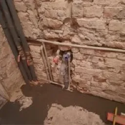 Surowa kamienna ściana z widocznymi rurami i świeżo wylaną, szarą posadzką samopoziomującą w trakcie remontu.