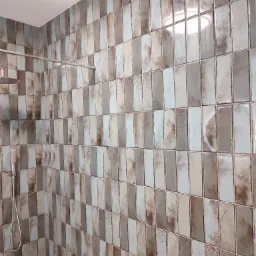 Ściana kabiny prysznicowej wyłożona podłużnymi, błyszczącymi płytkami w odcieniach beżu, brązu i błękitu, z widoczną deszczownicą i słuchawką prysznicową.