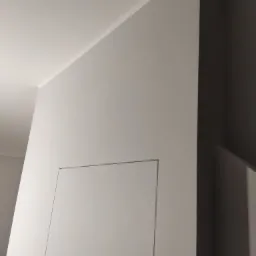 Szara ściana z ukrytymi drzwiami rewizyjnymi, widoczny fragment sufitu i narożnika, minimalistyczny design.