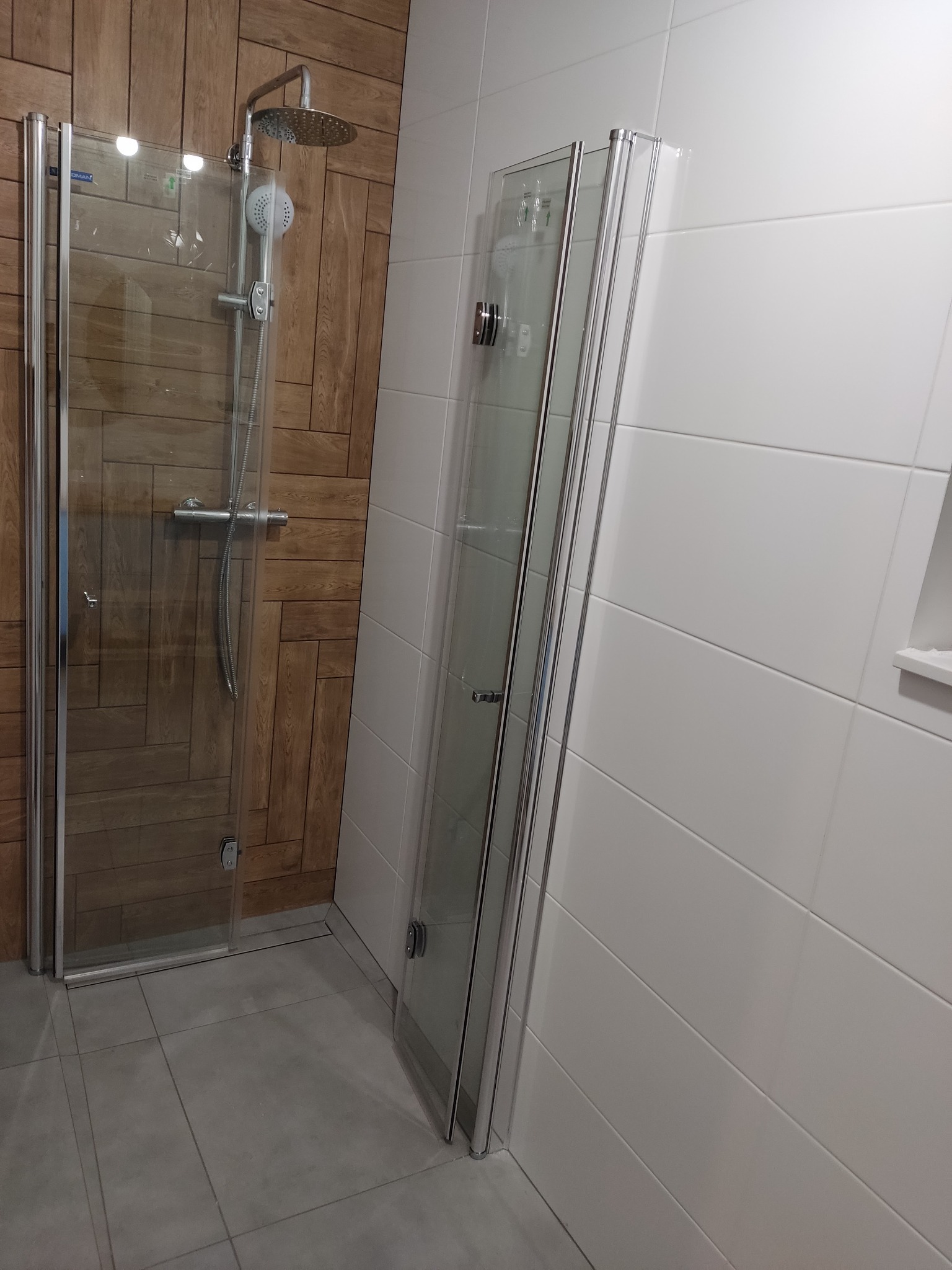 Zamontowana kabina prysznicowa z deszczownicą na tle drewnopodobnych płytek i białych płytek ściennych, widoczna szara podłoga.