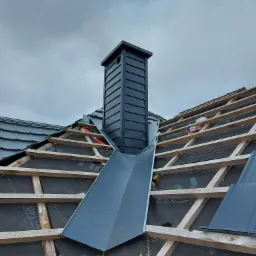 Montaż obróbek blacharskich przy kominie na dachu z drewnianą konstrukcją i membraną dachową, widoczne fragmenty dachówek i pochmurne niebo.