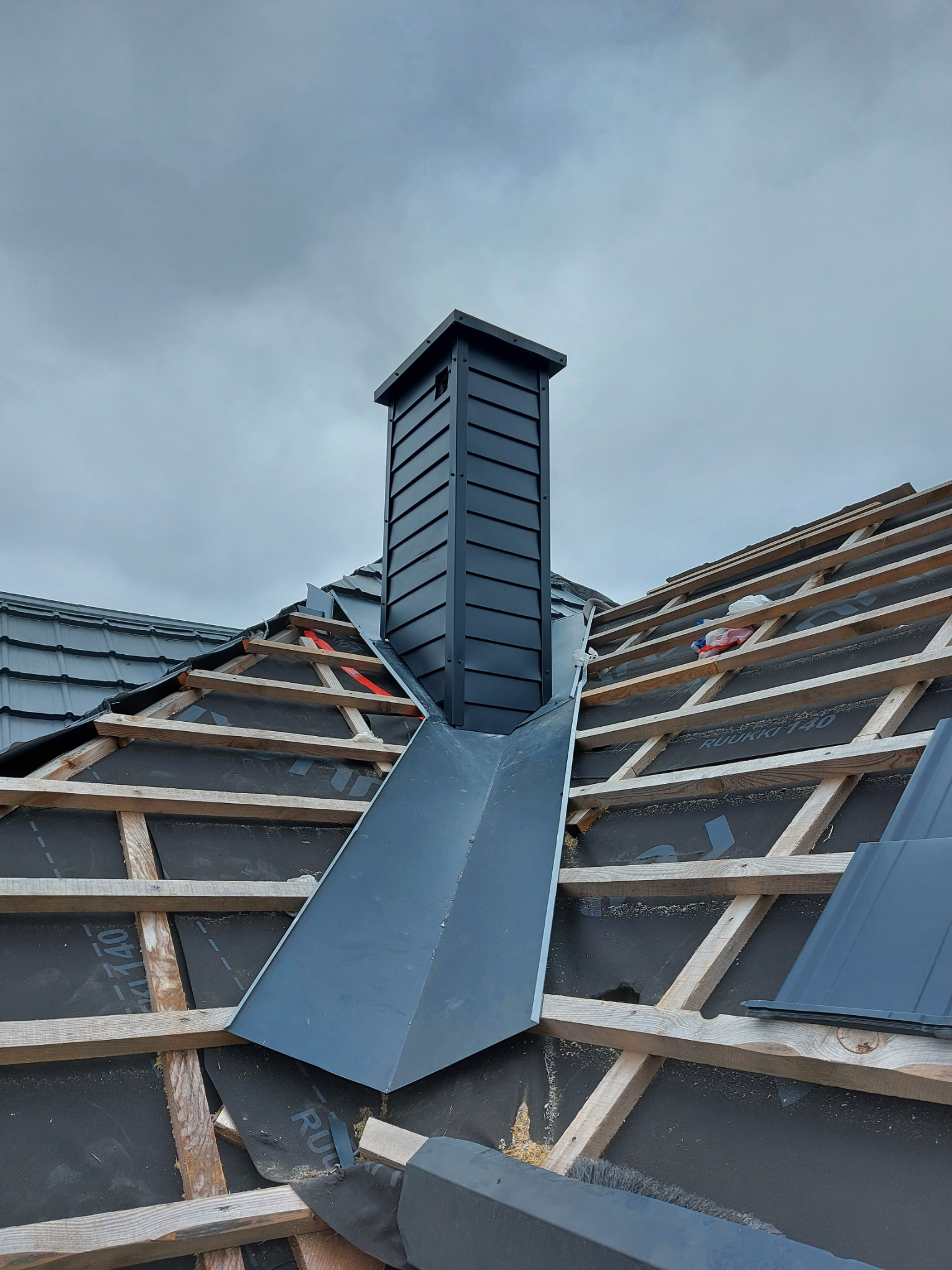 Montaż obróbek blacharskich przy kominie na dachu z drewnianą konstrukcją i membraną dachową, widoczne fragmenty dachówek i pochmurne niebo.