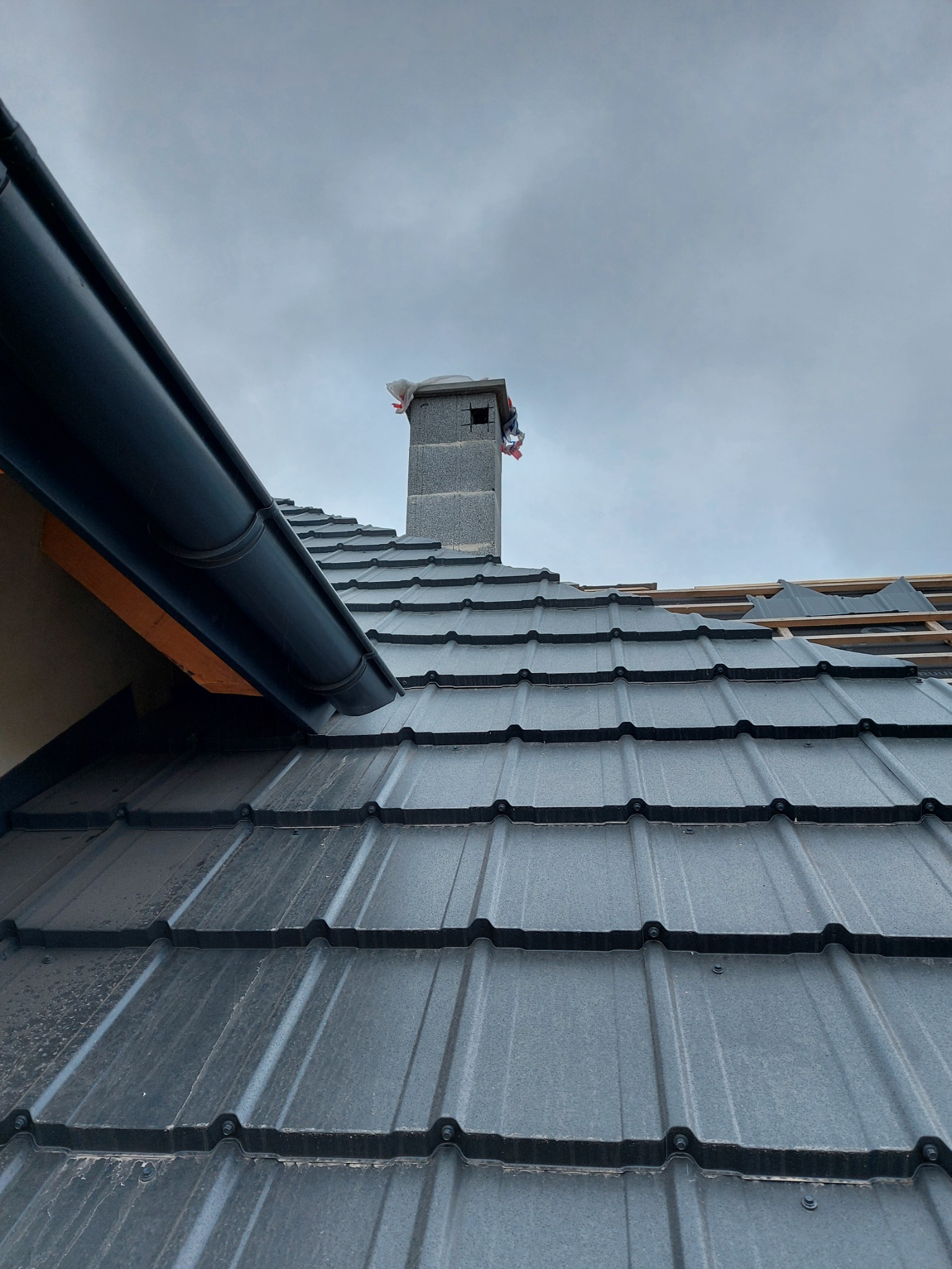Montaż czarnej blachodachówki modułowej na dachu domu w Łomży, widoczny komin z pustaków oraz fragment orynnowania.