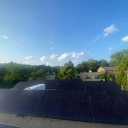 Liqene Fotowoltaika Radom, Pompy Ciepła - Panele słoneczne na dachu budynku w słoneczny dzień. Widoczne drzewa i zabudowa w tle. Biały element na dachu. Ujęcie z góry.