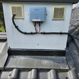 Liqene Fotowoltaika Radom, Pompy Ciepła - Szary moduł sterujący Inccy ERS zamontowany na kominie z dwoma kratkami wentylacyjnymi, czarne rury ochronne na przewody, dachówka ceramiczna.