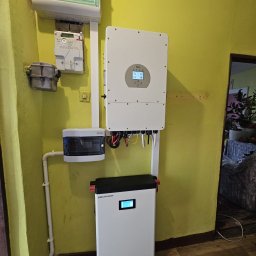 Liqene Fotowoltaika Radom, Pompy Ciepła - Zamontowany na żółtej ścianie magazyn energii Heckman z inwerterem Deye, skrzynką bezpiecznikową i licznikiem. Widoczne kable i rurki instalacyjne.