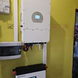 Liqene Fotowoltaika Radom, Pompy Ciepła - Zamontowany biały falownik Deye z podłączonymi kablami na żółtej ścianie, obok skrzynka elektryczna i biały akumulator Heckman. Widoczny licznik energii.