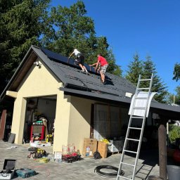 Liqene Fotowoltaika Radom, Pompy Ciepła - Montaż paneli na dachu garażu przez dwóch instalatorów w słoneczny dzień. Widoczna drabina i narzędzia. Ujęcie z dołu, zieleń drzew w tle.