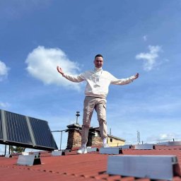 Liqene Fotowoltaika Radom, Pompy Ciepła - Mężczyzna stoi na dachu z panelami słonecznymi, rozkładając ręce na tle błękitnego nieba z chmurami. Widoczne elementy montażowe paneli.