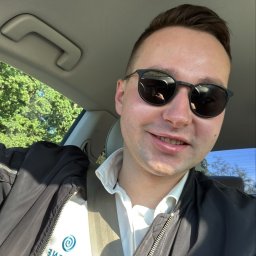 Liqene Fotowoltaika Radom, Pompy Ciepła - Uśmiechnięty mężczyzna w okularach przeciwsłonecznych w samochodzie, z widocznym logo firmy na koszuli. Widok z perspektywy kierowcy, drzewa w tle.