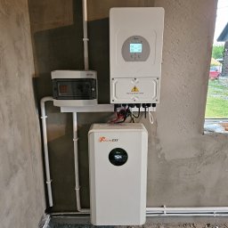 Liqene Fotowoltaika Radom, Pompy Ciepła - Zestaw urządzeń: falownik Daye, Felicity ESS i rozdzielnica ETI, połączone białymi rurami PCV na surowej ścianie. Widoczne okablowanie i okno w tle.