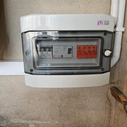 Liqene Fotowoltaika Radom, Pompy Ciepła - Zamknięta skrzynka elektryczna ETI z bezpiecznikami i ogranicznikami przepięć, zamontowana na szarej ścianie. Widoczne przewody i rury instalacyjne.