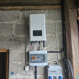 Liqene Fotowoltaika Radom, Pompy Ciepła - Zamontowany falownik Deye na szarej ścianie z betonu komórkowego. Widoczne przewody, skrzynka z bezpiecznikami i gniazda siłowe. Surowy, techniczny charakter wnętrza.