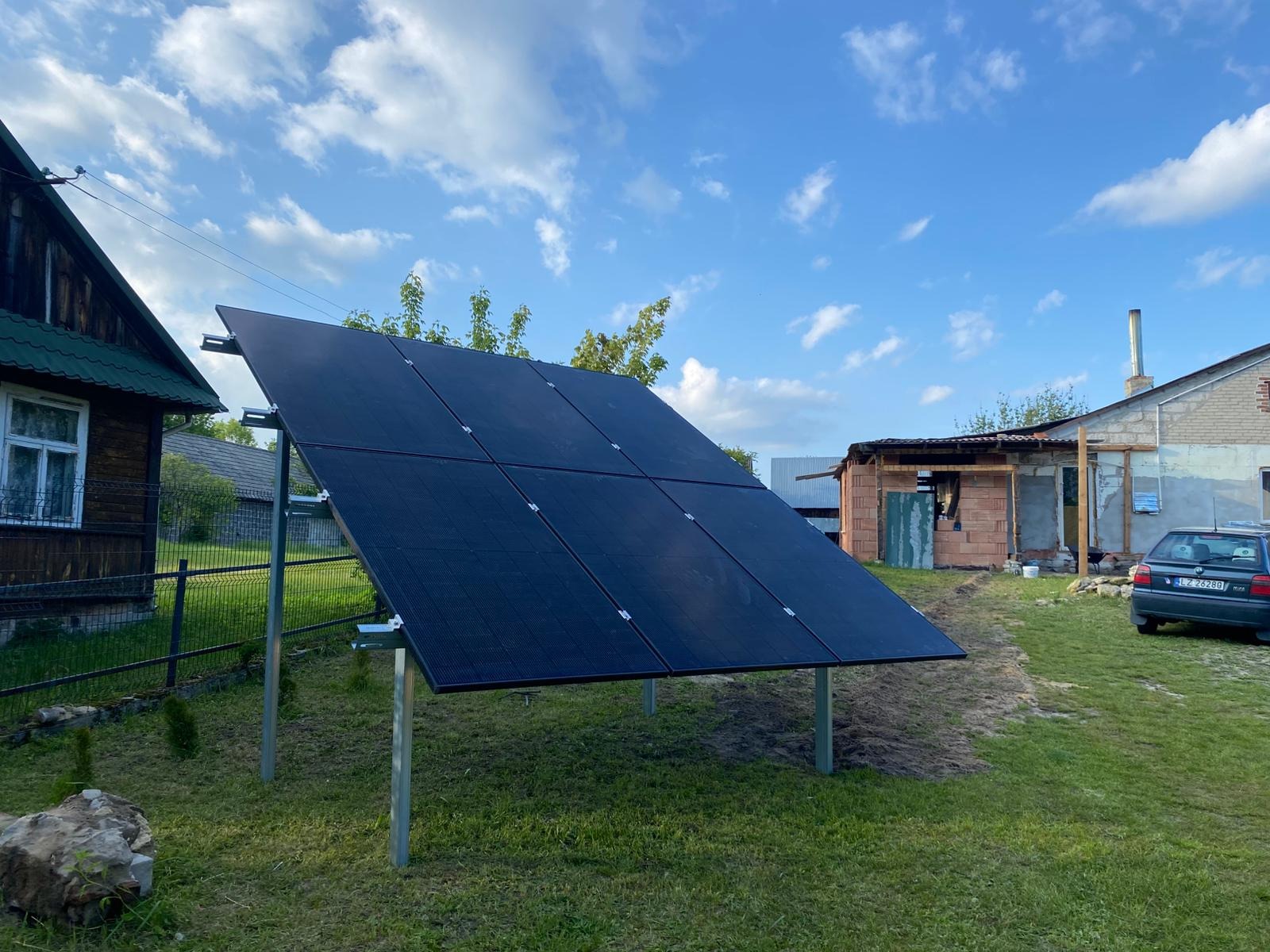 Panel fotowoltaiczny na metalowej konstrukcji, ustawiony na trawniku przed domem z cegły w słoneczny dzień. Widoczne błękitne niebo z chmurami.