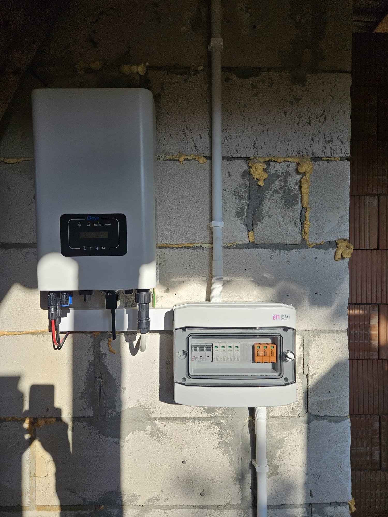 Zamontowany falownik fotowoltaiczny marki Deye z podłączonymi przewodami i skrzynką bezpiecznikową na szarej ścianie z betonu komórkowego.