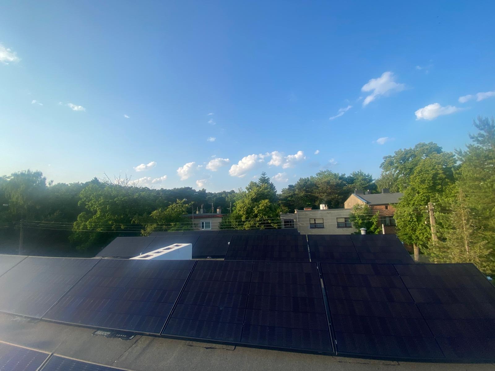 Panele słoneczne na dachu budynku w słoneczny dzień. Widoczne drzewa i zabudowa w tle. Biały element na dachu. Ujęcie z góry.