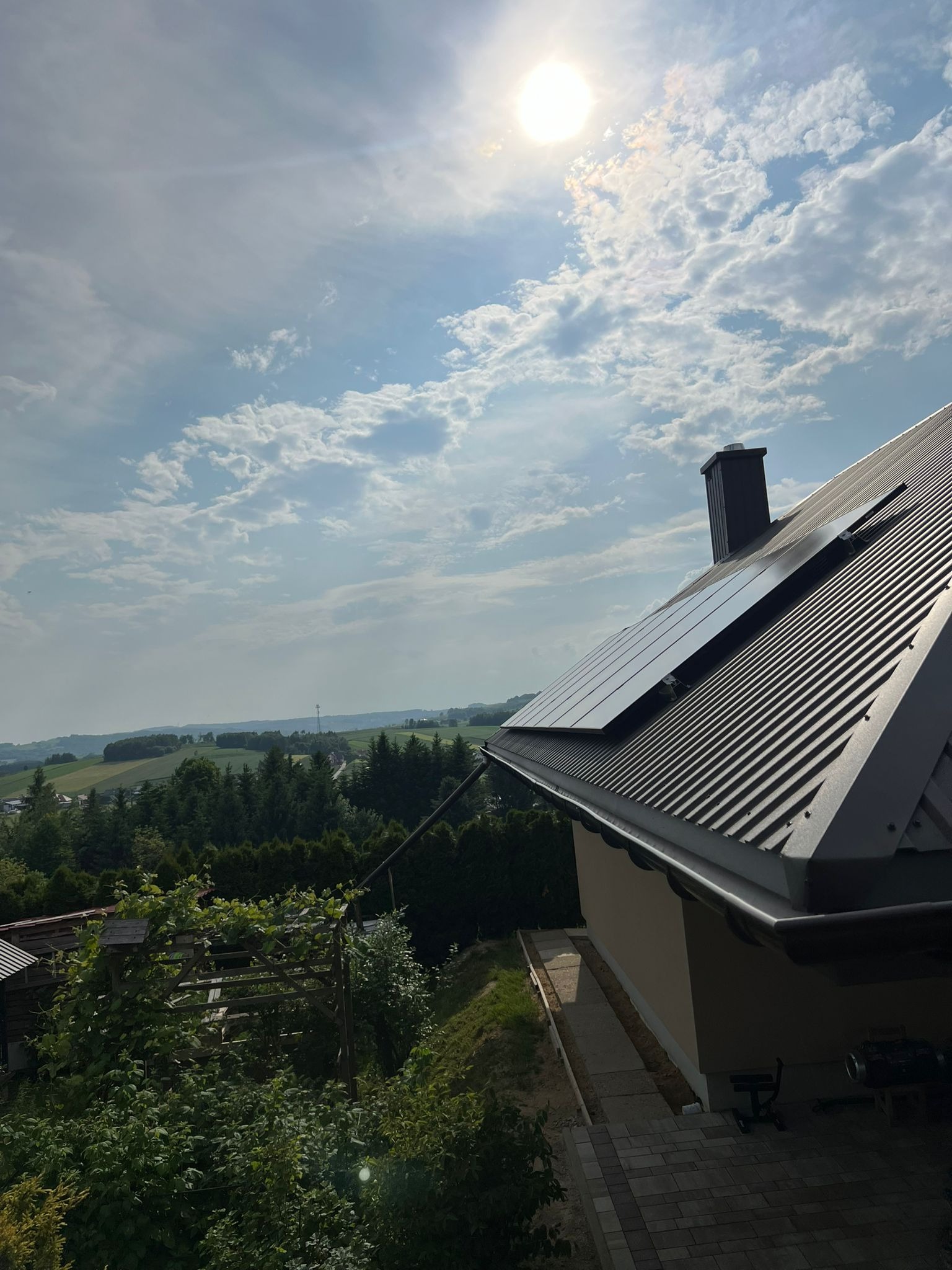 Panele słoneczne na dachu domu z ciemnej blachy, widok na zielone wzgórza pod błękitnym, zachmurzonym niebem z jasnym słońcem. Ujęcie z dołu.