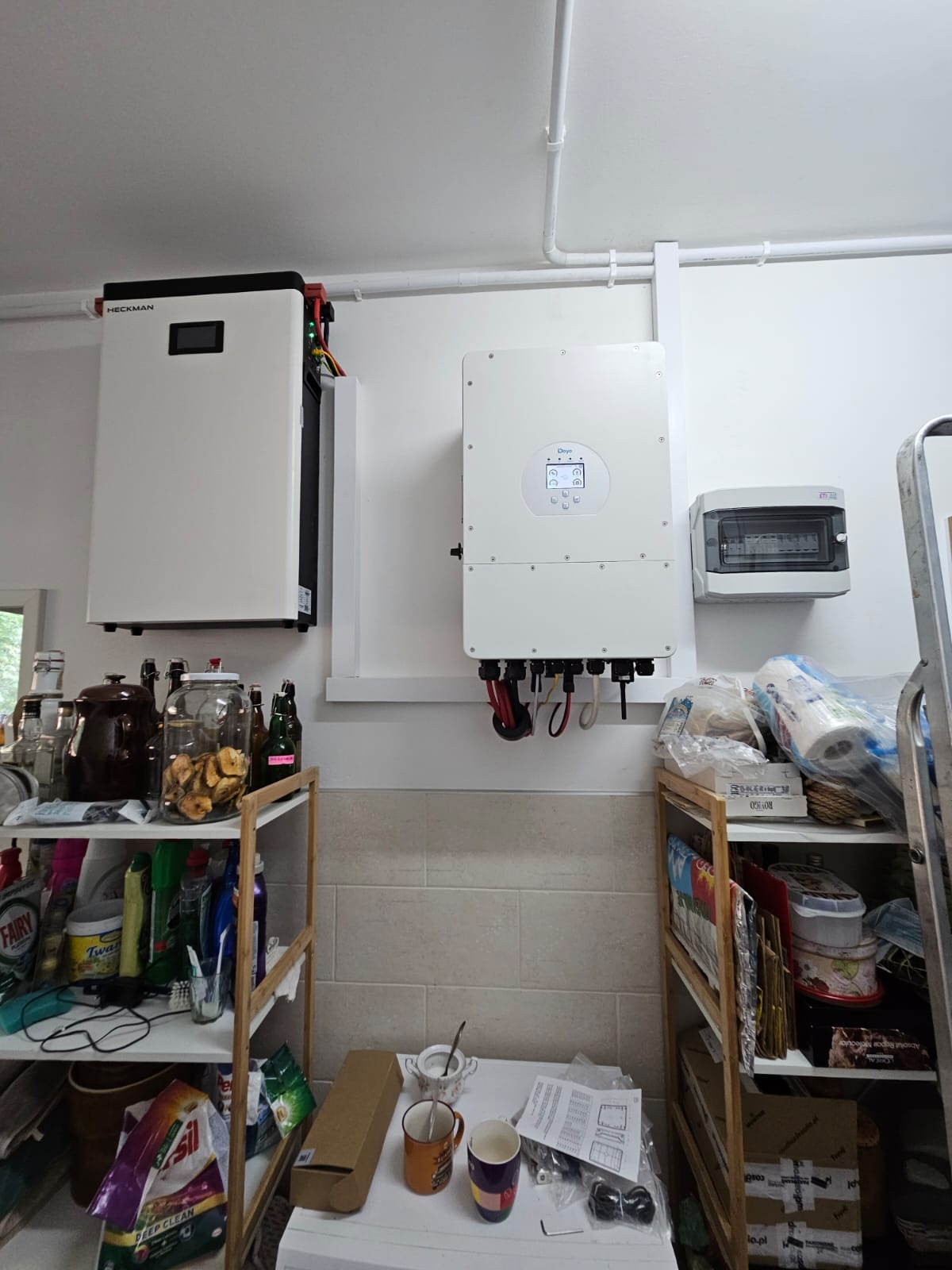 Biały falownik i magazyn energii Heckman zamontowane na ścianie w pomieszczeniu. Widoczne kable i przewody. Poniżej półki z różnymi przedmiotami.