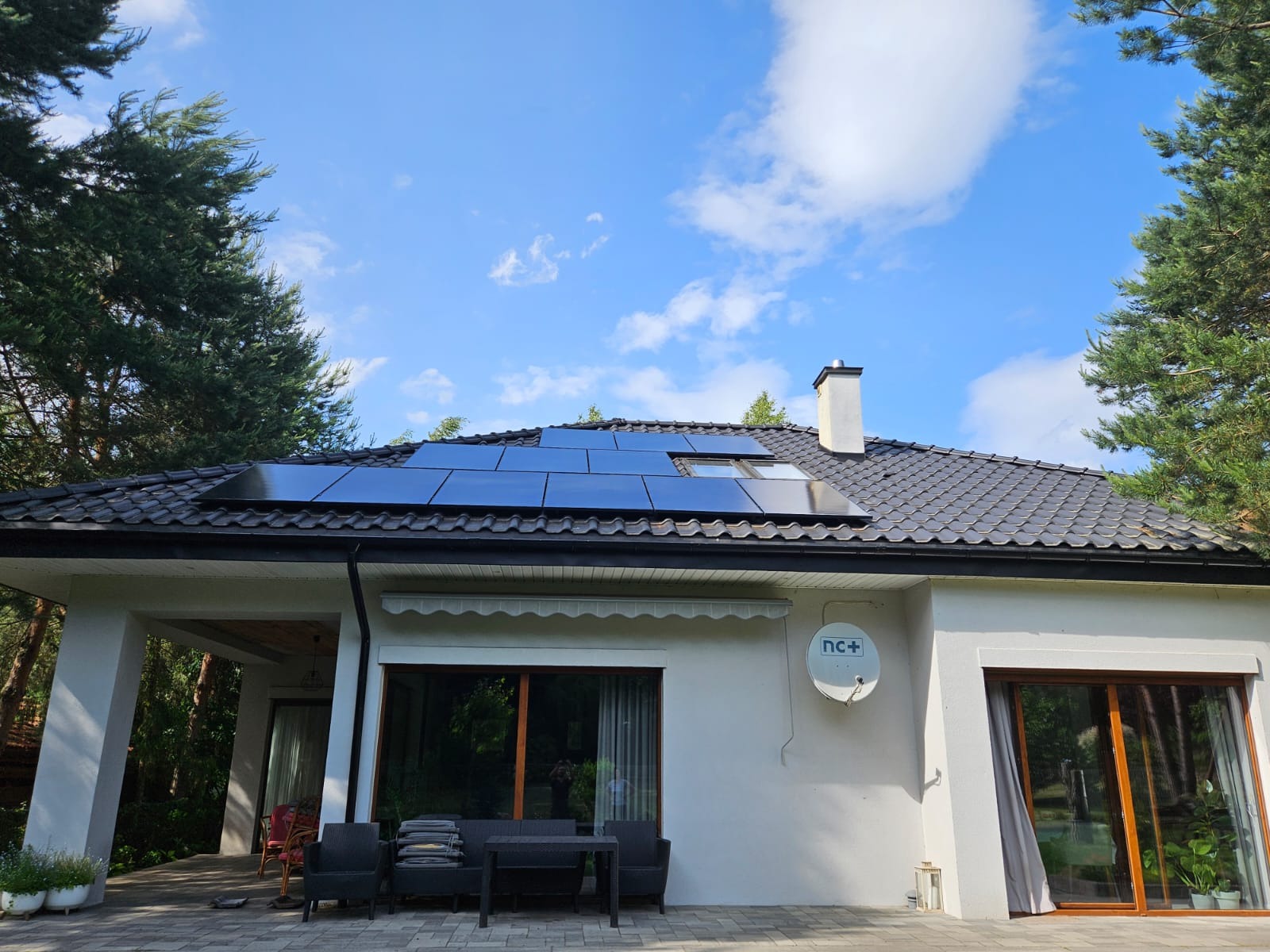 Dom z panelami fotowoltaicznymi na ciemnym dachu, widok z dołu. Biała elewacja, okna i antena satelitarna. Drzewa po bokach kadru i błękitne niebo.