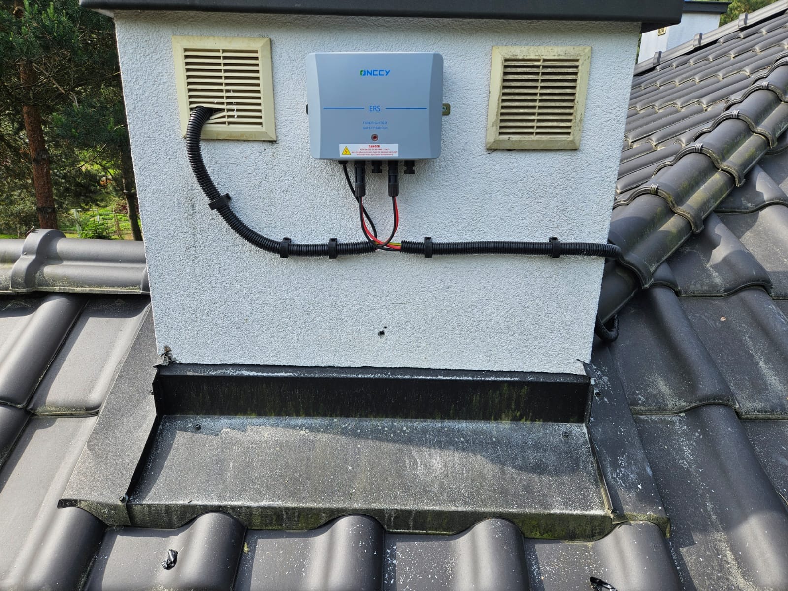 Szary moduł sterujący Inccy ERS zamontowany na kominie z dwoma kratkami wentylacyjnymi, czarne rury ochronne na przewody, dachówka ceramiczna.