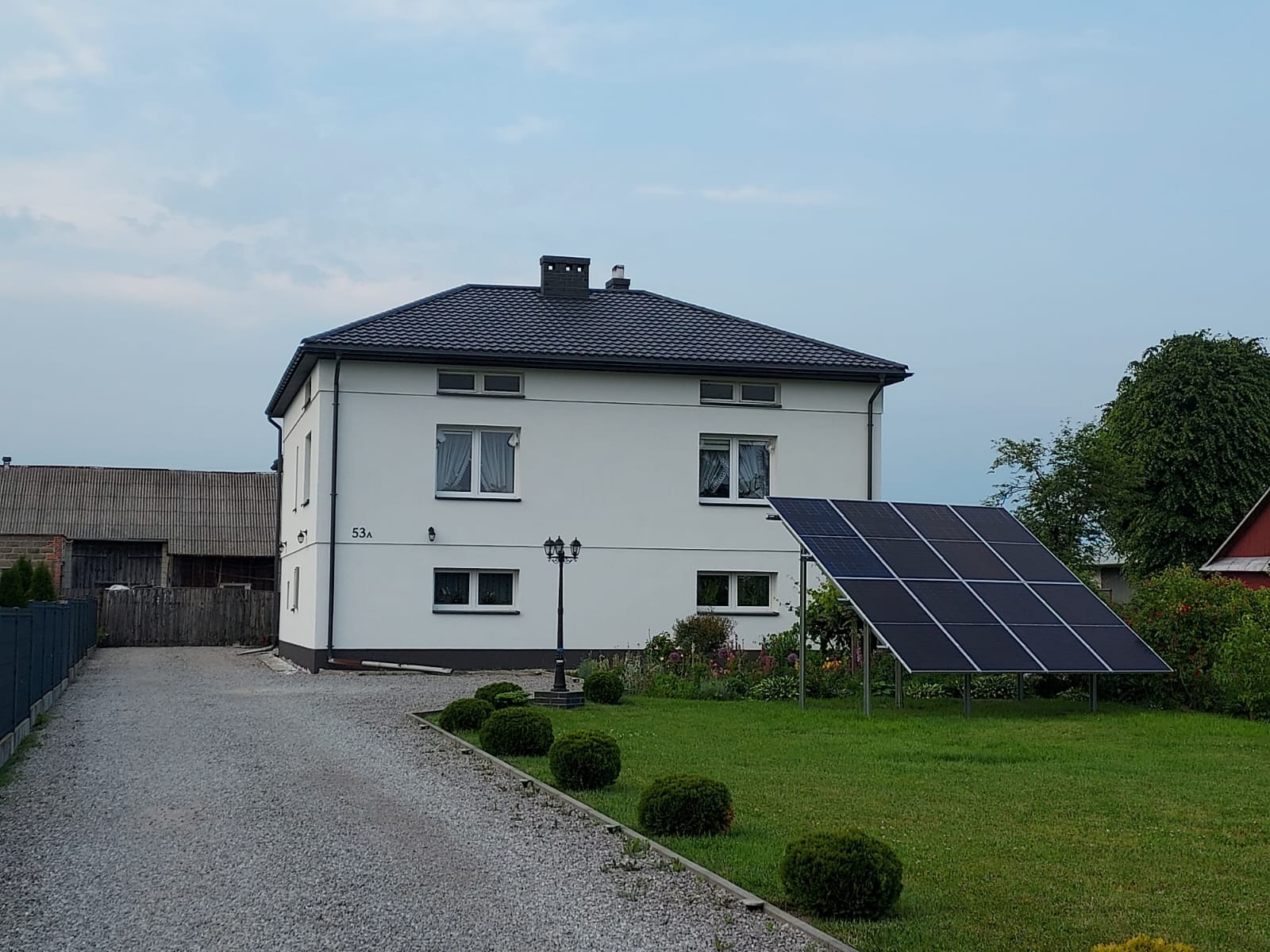 Dom z panelami fotowoltaicznymi na trawniku w Radomiu. Instalacja wolnostojąca, widok z dołu, w tle budynek mieszkalny z czarnym dachem i białymi ścianami.