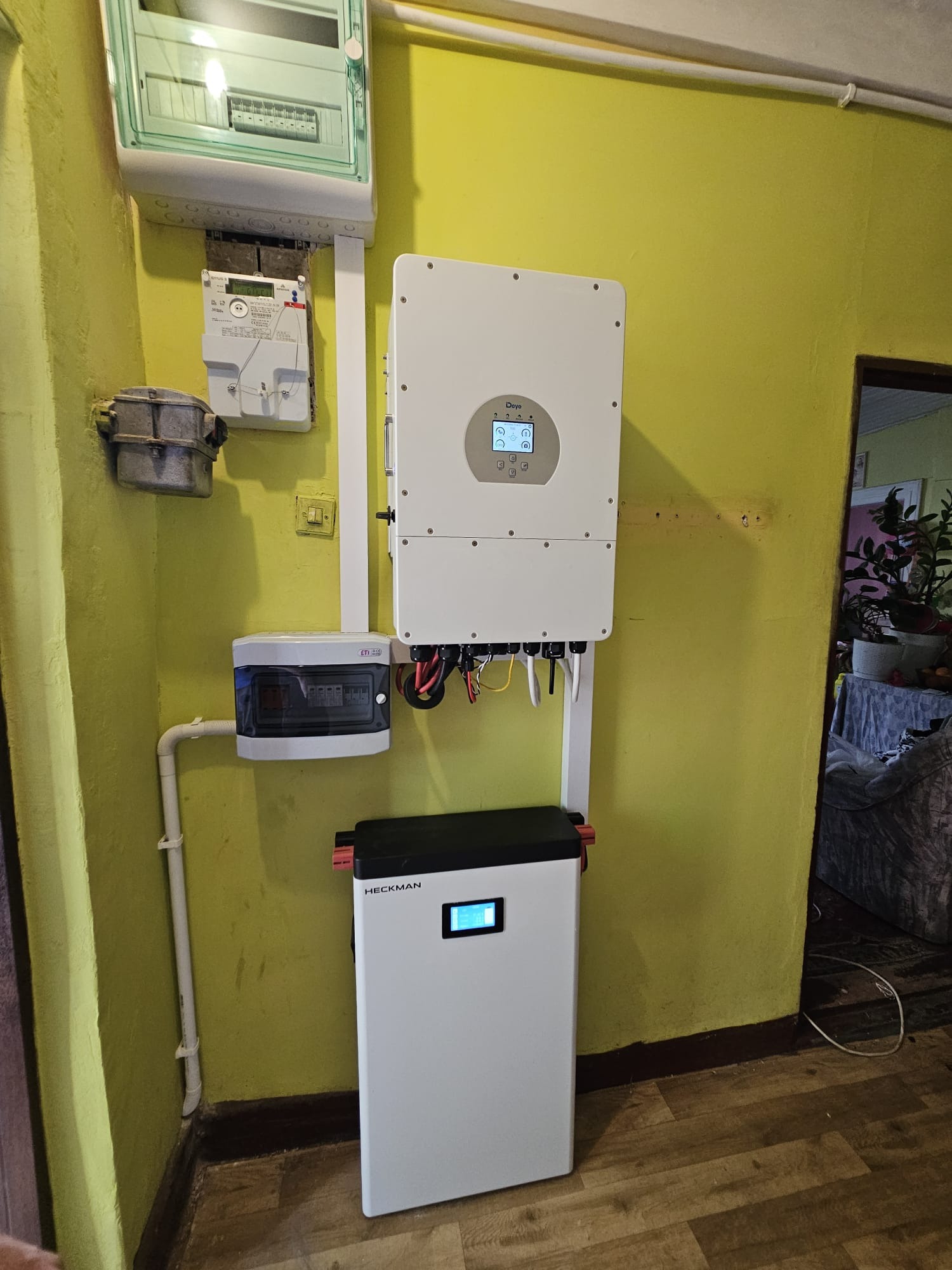 Zamontowany na żółtej ścianie magazyn energii Heckman z inwerterem Deye, skrzynką bezpiecznikową i licznikiem. Widoczne kable i rurki instalacyjne.