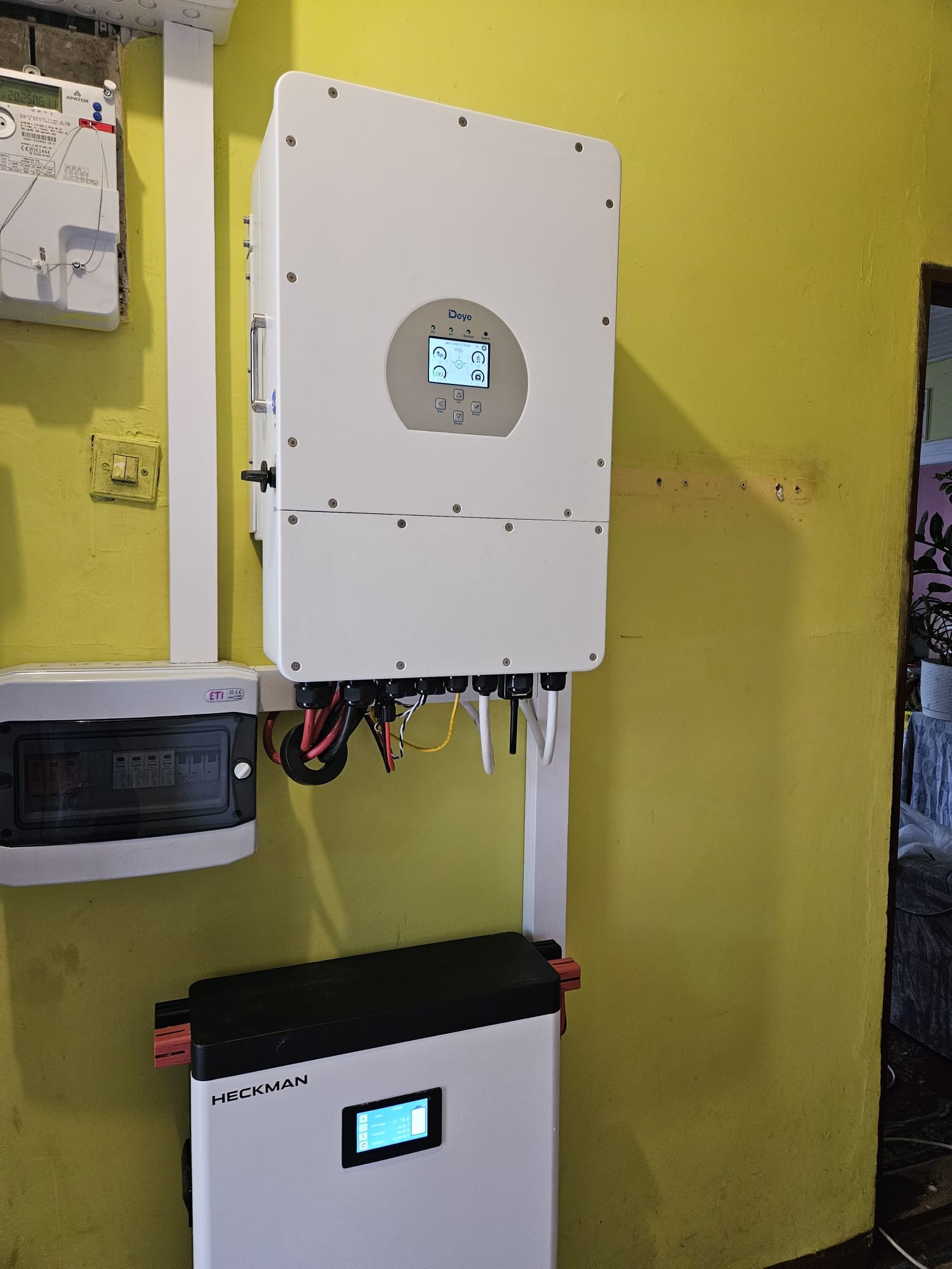 Zamontowany biały falownik Deye z podłączonymi kablami na żółtej ścianie, obok skrzynka elektryczna i biały akumulator Heckman. Widoczny licznik energii.