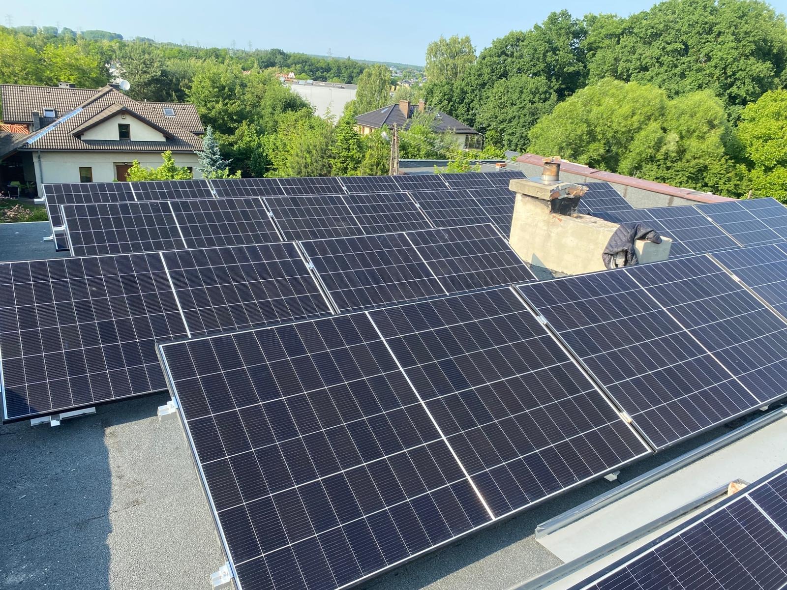 Widok z góry na zainstalowane panele słoneczne na dachu budynku z kominem, otoczonego zielenią drzew. W tle widoczne zabudowania.
