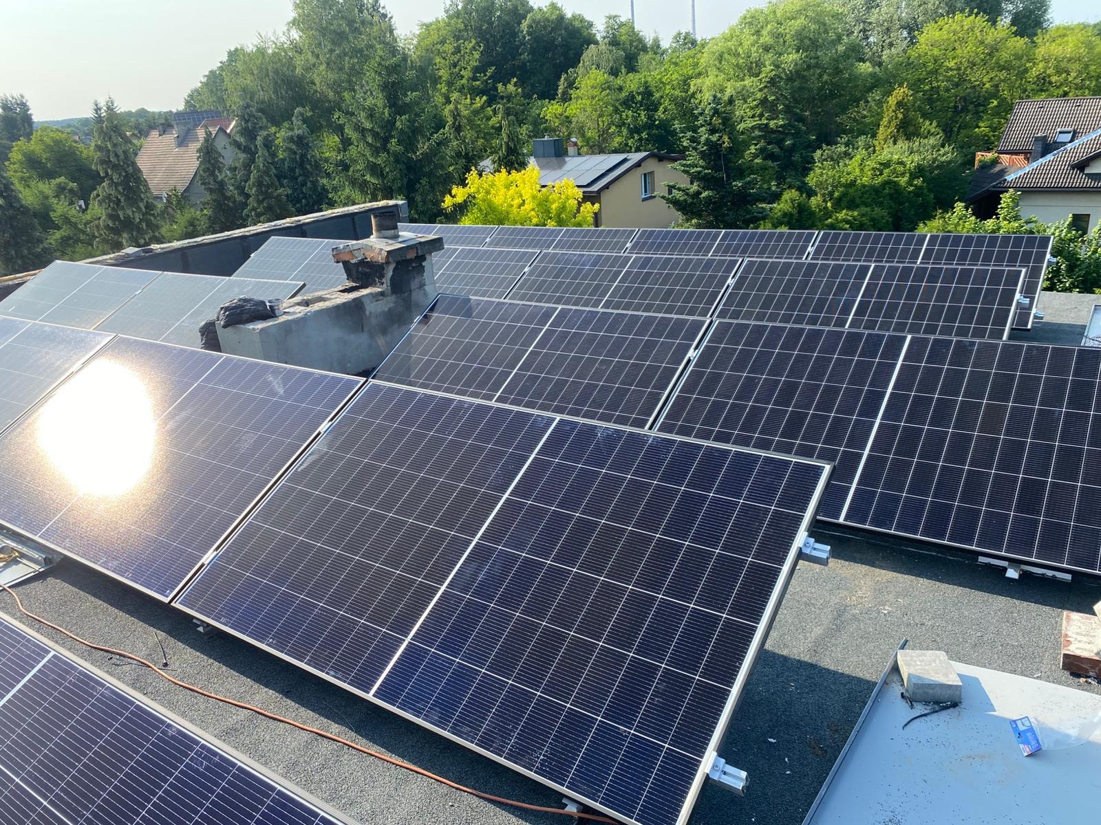 Fotowoltaika na dachu domu jednorodzinnego, widok z góry. Panele w rzędach, komin w tle, zieleń drzew w tle. Słoneczne odbicie na jednym z paneli.