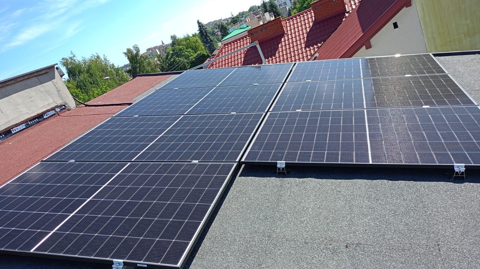 Panele fotowoltaiczne na dachu pokrytym papą, z widocznymi elementami montażowymi i fragmentem czerwonej dachówki w tle, w słoneczny dzień.
