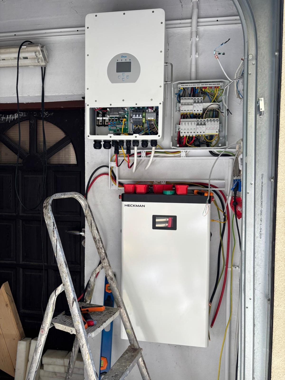 Falownik Deye, akumulator Heckman, rozdzielnica i drabina monterska. Instalacja w garażu, widoczne kable i przewody elektryczne. Elementy instalacji fotowoltaicznej.