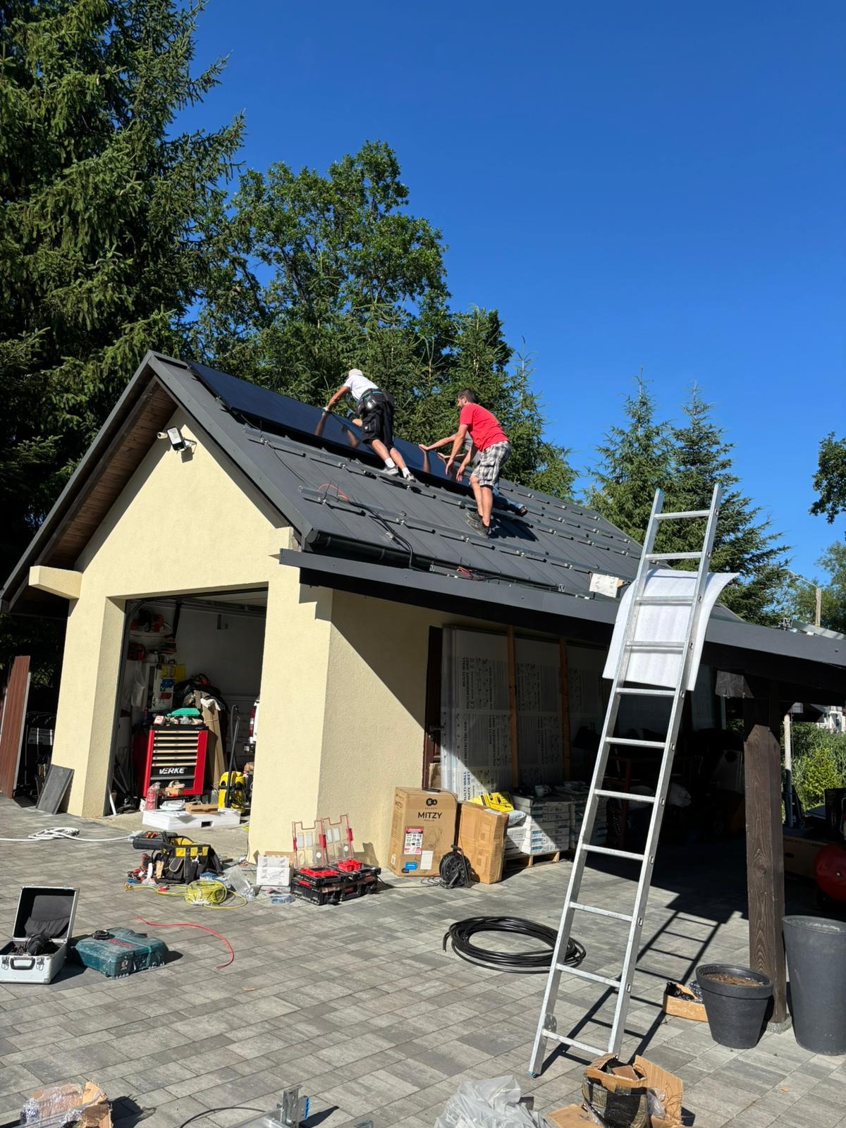Montaż paneli na dachu garażu przez dwóch instalatorów w słoneczny dzień. Widoczna drabina i narzędzia. Ujęcie z dołu, zieleń drzew w tle.