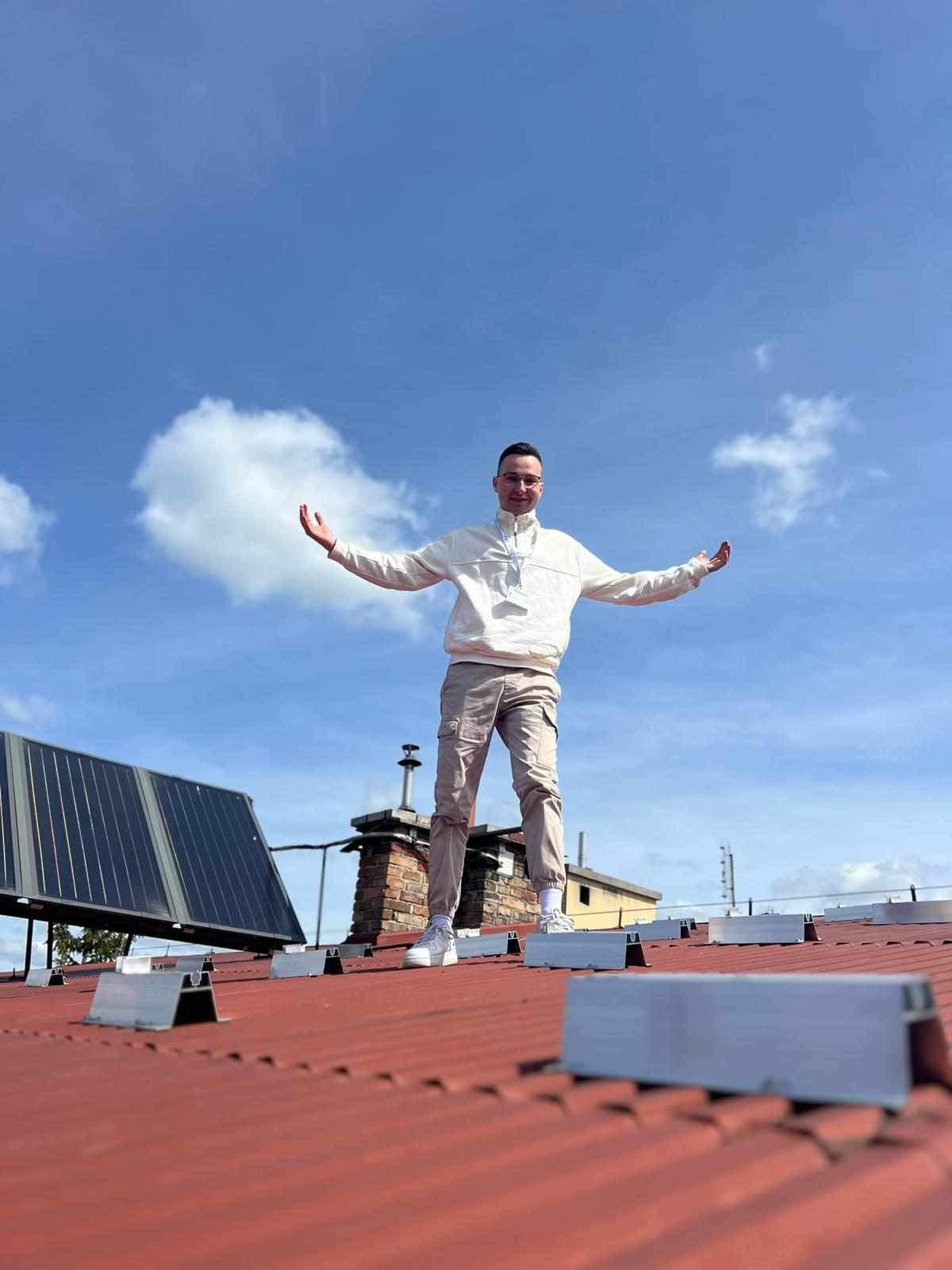 Mężczyzna stoi na dachu z panelami słonecznymi, rozkładając ręce na tle błękitnego nieba z chmurami. Widoczne elementy montażowe paneli.