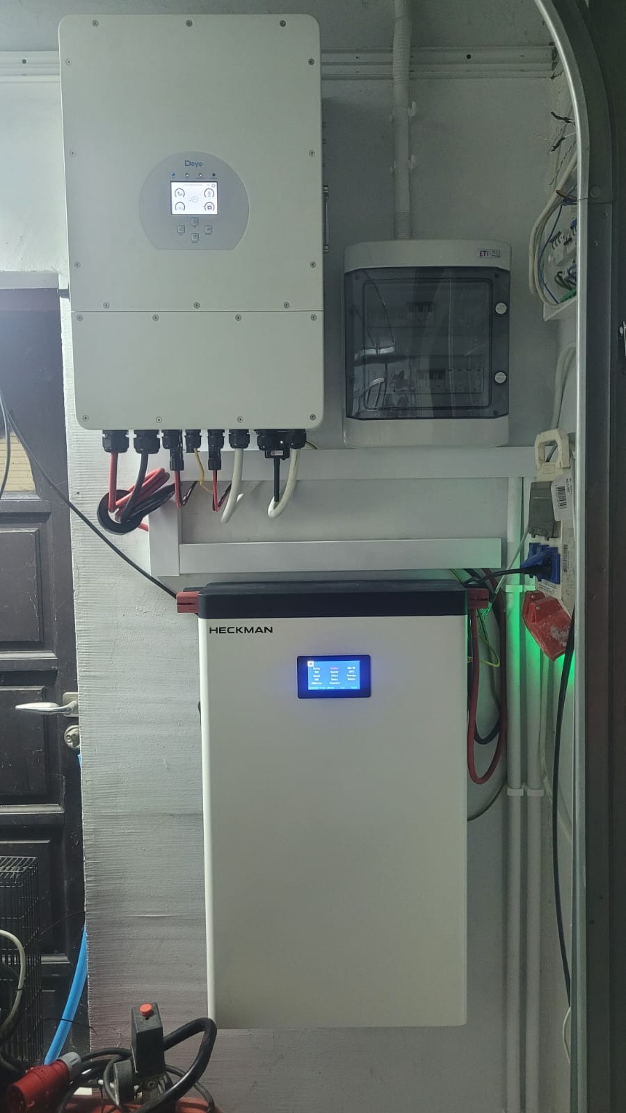Zamontowany magazyn energii Heckman z wyświetlaczem LCD, obok skrzynka elektryczna i falownik Deye. Widoczne kable i przewody. Całość zamontowana na ścianie wewnątrz budynku.