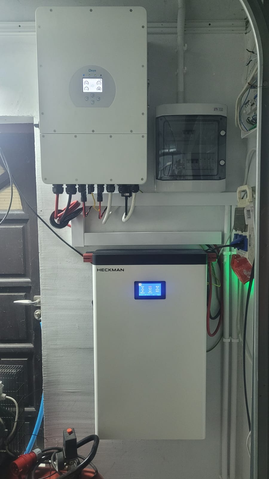 Zamontowany inwerter Deye i magazyn energii Heckman z wyświetlaczem, obok skrzynka elektryczna. Widoczne kable i przewody. Instalacja wewnątrz budynku.