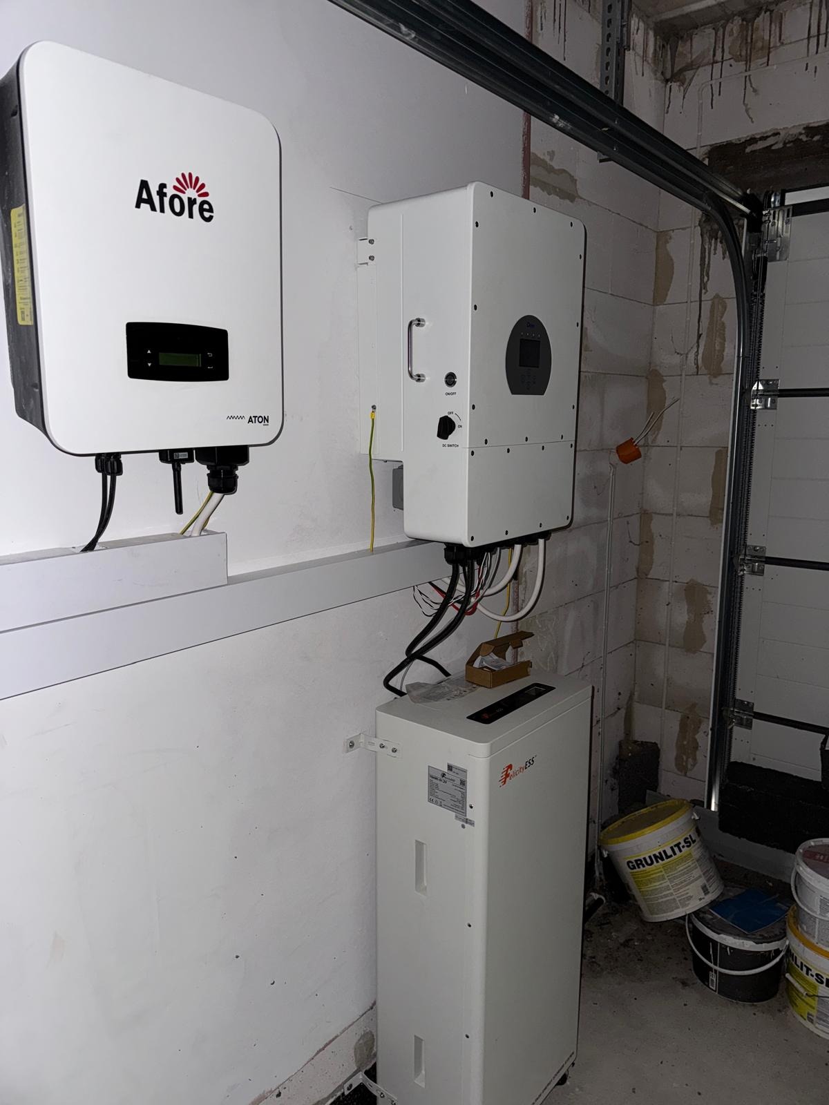 Zamontowane falowniki Afore i Eliness ESS w garażu, widoczne kable i puszki instalacyjne. Białe ściany, brama garażowa. Instalacja fotowoltaiczna wewnątrz budynku.