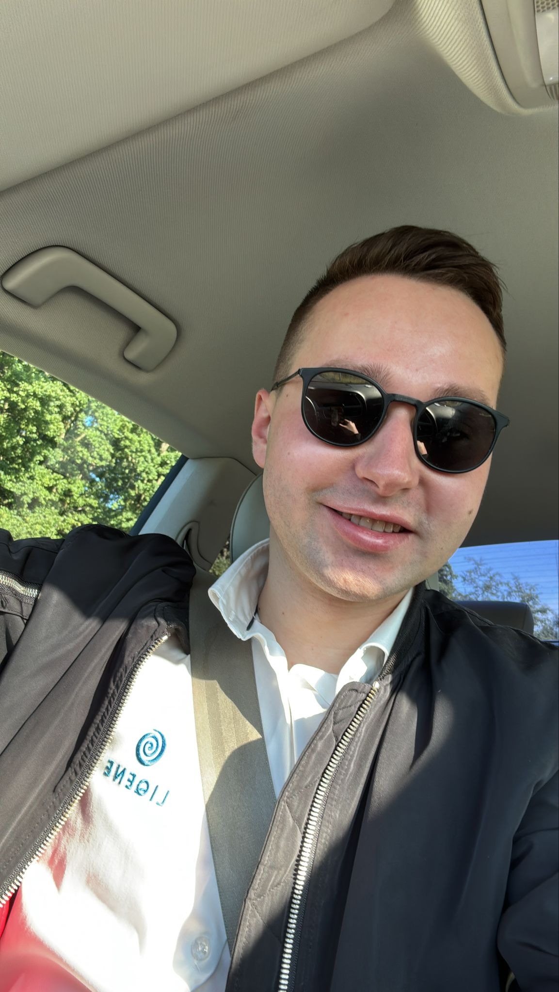 Uśmiechnięty mężczyzna w okularach przeciwsłonecznych w samochodzie, z widocznym logo firmy na koszuli. Widok z perspektywy kierowcy, drzewa w tle.