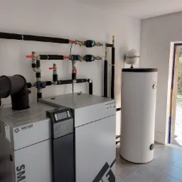 Nowoczesny piec CO marki Metal-Fach z zasobnikiem ciepłej wody użytkowej w kotłowni, instalacja grzewcza z czarnymi rurami izolowanymi i czerwonymi zaworami, widok z boku na otwarte drzwi.