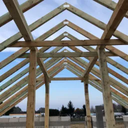 Konstrukcja drewniana dachu w trakcie budowy, widok od wewnątrz, z widocznymi elementami mocującymi i pustakami ceramicznymi w dolnej części kadru, na tle błękitnego nieba.