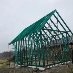 Szkielet drewnianej konstrukcji domu w trakcie budowy, pomalowany na zielono, widoczny na tle szarego nieba i częściowo wykończonego budynku z ciemną dachówką, z rusztowaniami i fragmentem osoby po...