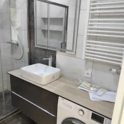 Nowoczesna łazienka po remoncie: biała umywalka nablatowa, bateria, podświetlane lustro z szafką, grzejnik drabinkowy, kabina prysznicowa i pralka Beko z dokumentacją techniczną.