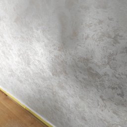 Alex-Bud - Ściana z dekoracyjną, srebrzystą fakturą przypominającą delikatne pociągnięcia pędzlem; widoczne zabezpieczenie taśmą malarską przy podłodze z drewnianych paneli.