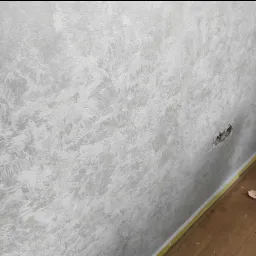 Ściana po nałożeniu dekoracyjnej, srebrzystej farby strukturalnej z widocznym gniazdkiem elektrycznym w trakcie montażu i taśmą malarską zabezpieczającą podłogę.