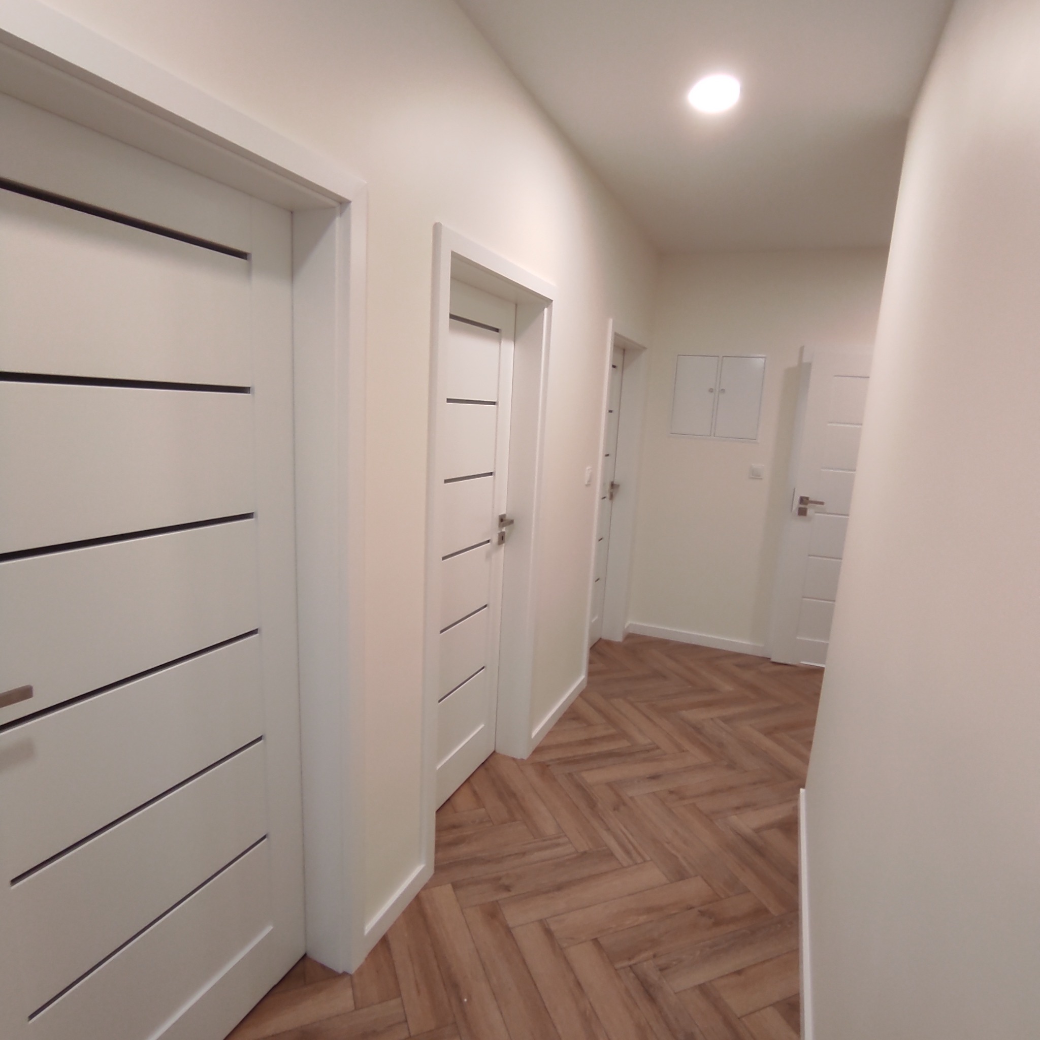 Korytarz z trzema białymi drzwiami z poziomymi, ciemnymi wstawkami i podłogą ułożoną w jodełkę z paneli imitujących drewno.