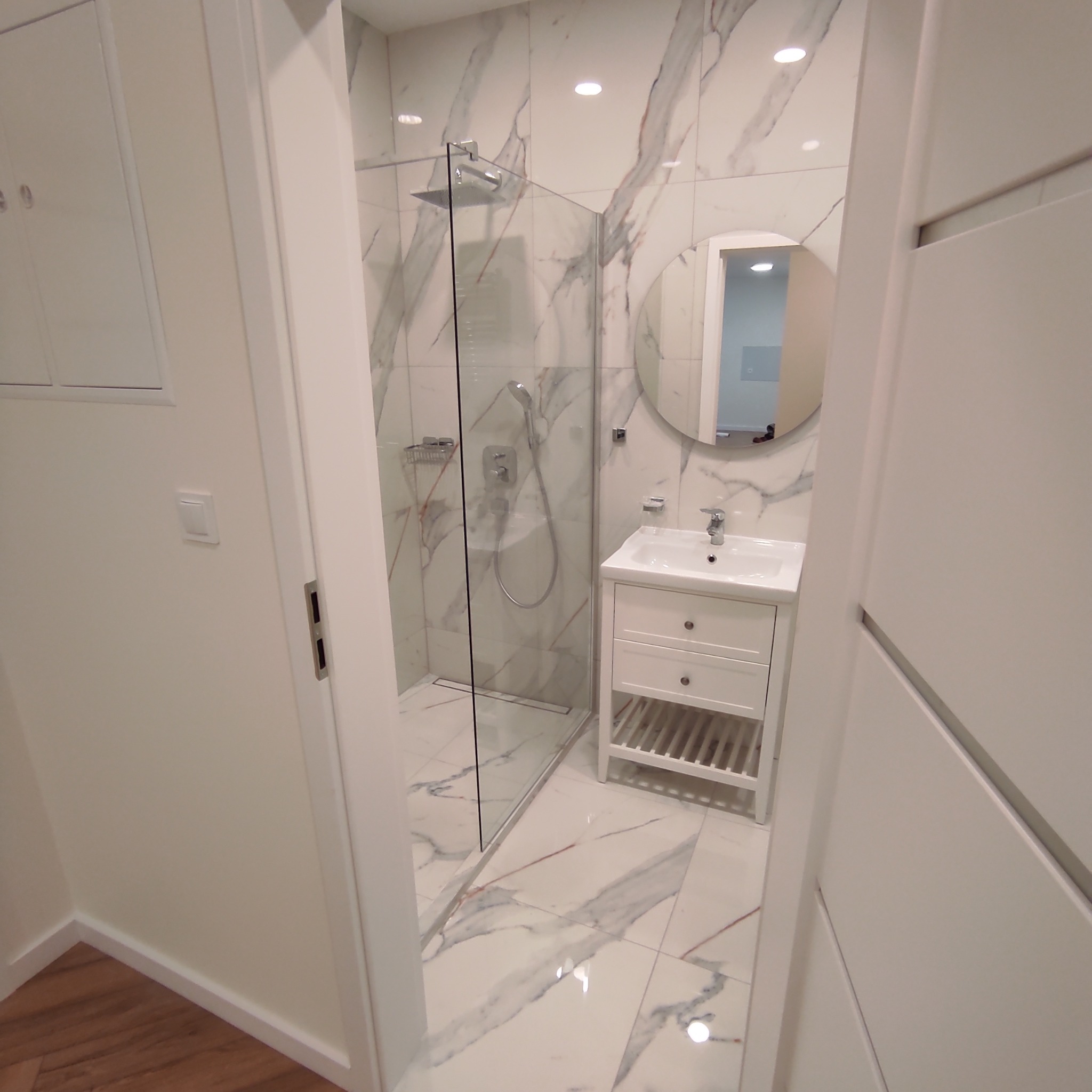 Nowoczesna łazienka z kabiną prysznicową typu walk-in, umywalką na szafce z szufladami i okrągłym lustrem, wykończona płytkami imitującymi marmur z wyraźnym żyłowaniem.