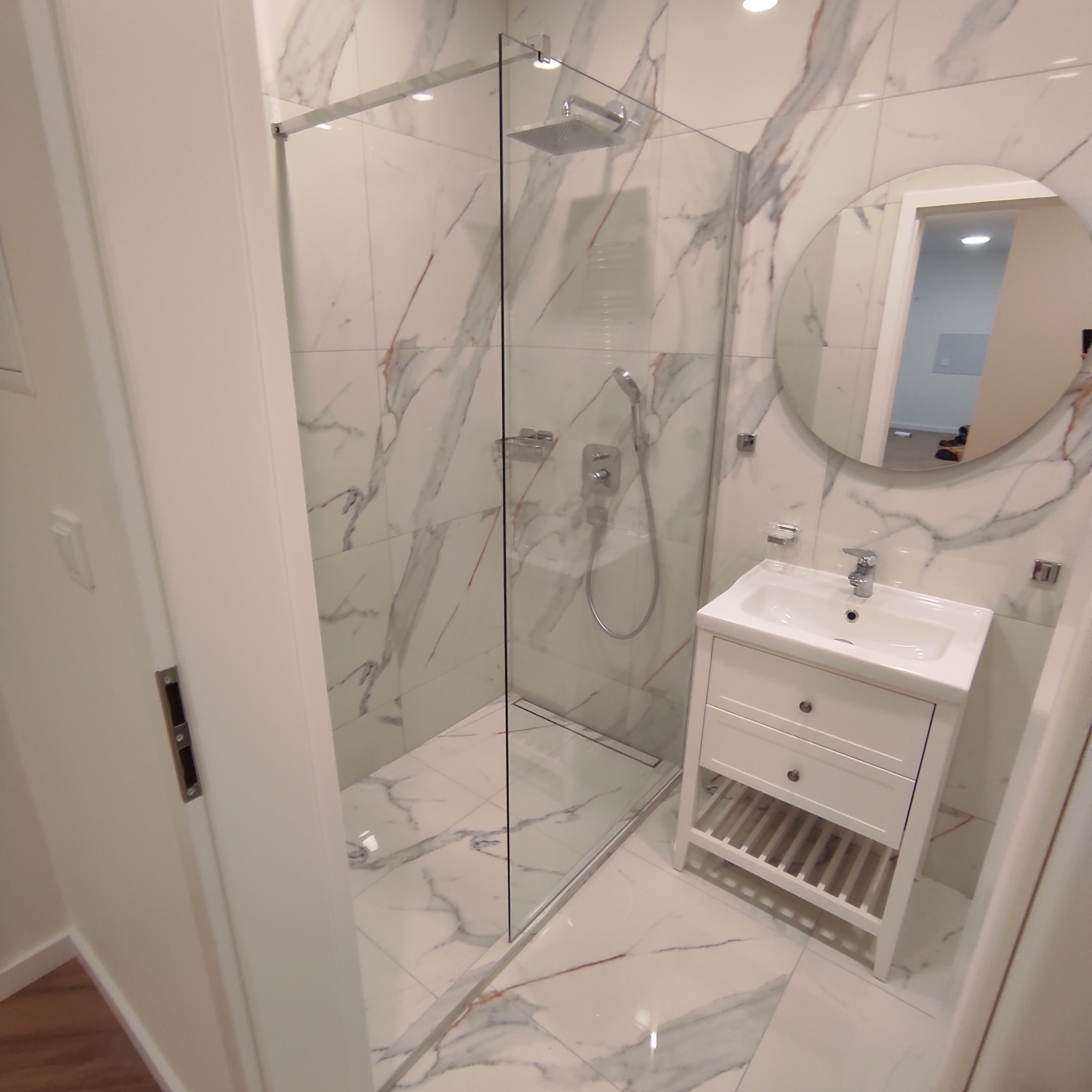 Wykończona łazienka z kabiną prysznicową typu walk-in, umywalką w białej szafce z szufladami i okrągłym lustrem, całość wykończona białymi płytkami imitującymi marmur.