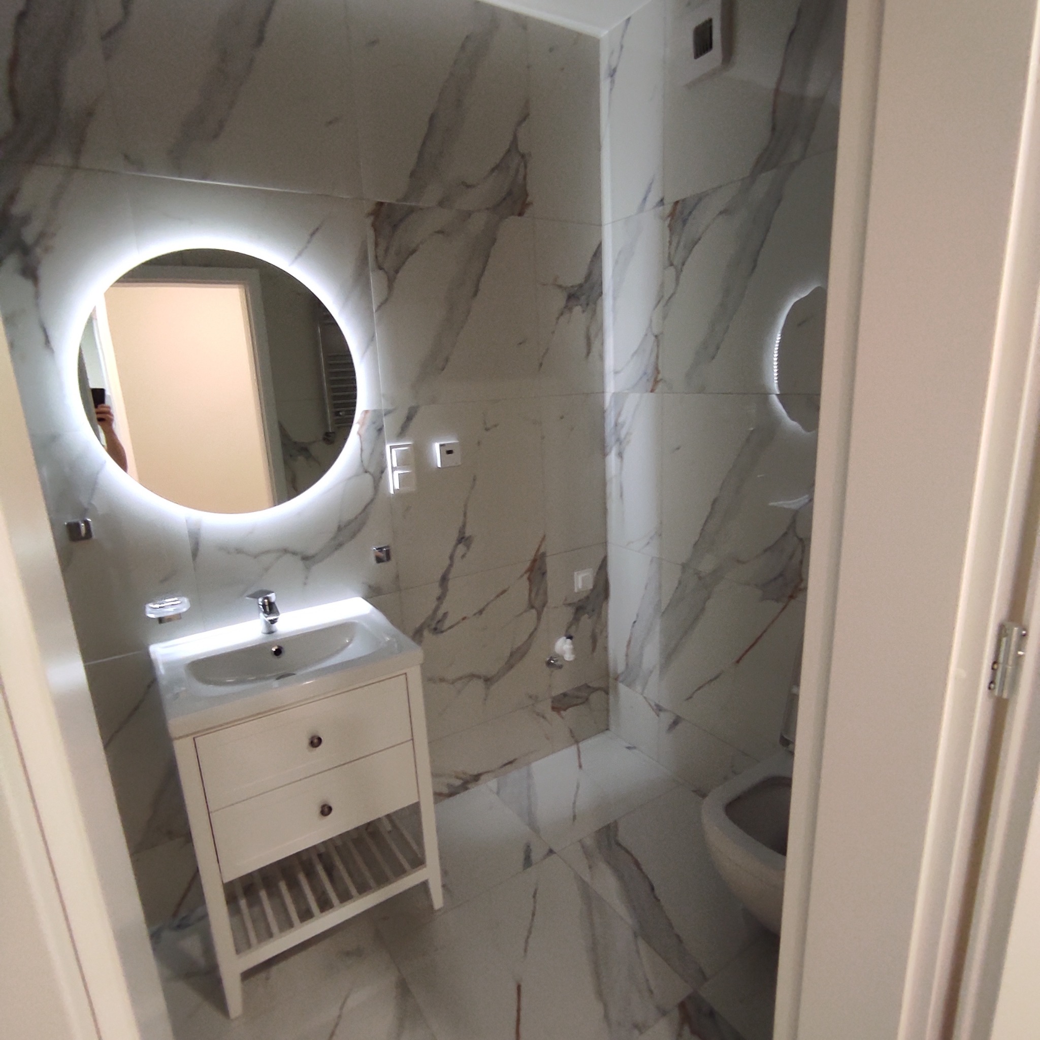 Elegancka łazienka z marmurowymi płytkami, okrągłym lustrem z podświetleniem LED i białą szafką umywalkową z szufladami i półką, widok zza uchylonych drzwi.