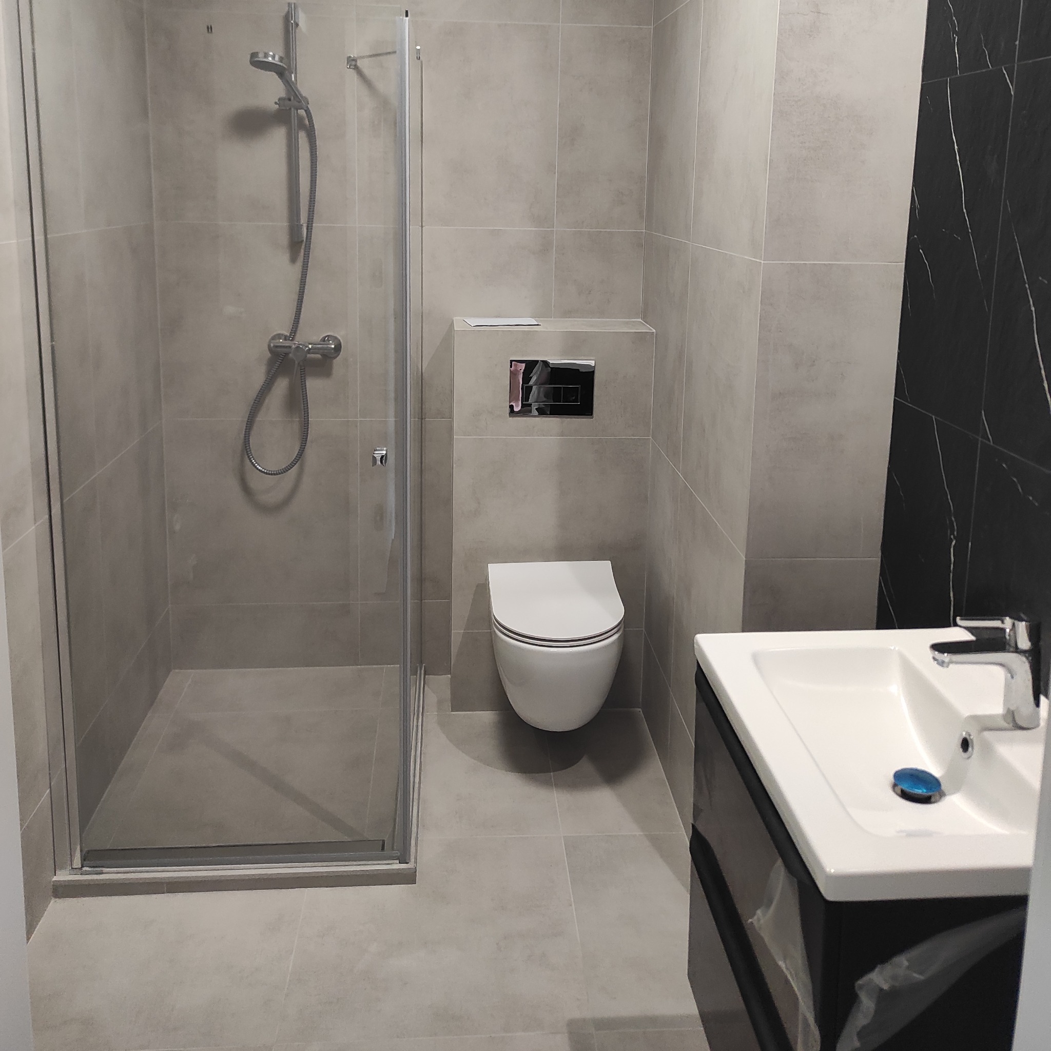 Nowoczesna łazienka w szarościach z akcentem czarnego marmuru, kabina prysznicowa ze szklanymi drzwiami, podwieszana toaleta z minimalistycznym przyciskiem, umywalka z szafką i chromowaną baterią.