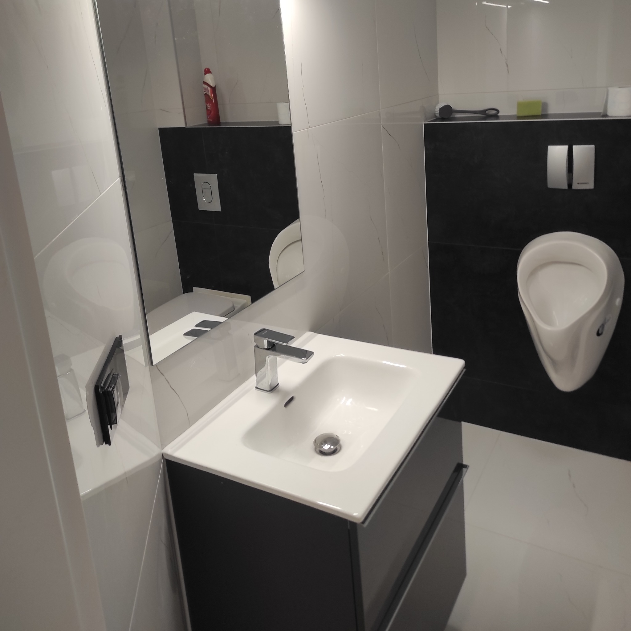 Nowoczesna łazienka z białymi płytkami imitującymi marmur, czarnymi akcentami i minimalistyczną umywalką z szafką w odcieniach szarości, widoczny pisuar i lustro.